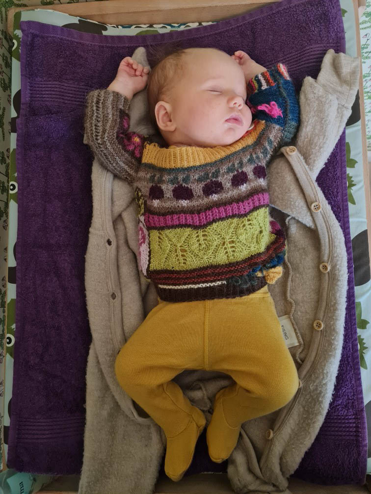 baby irma sleeping on back, wearing finished sweater, arms stretched over head